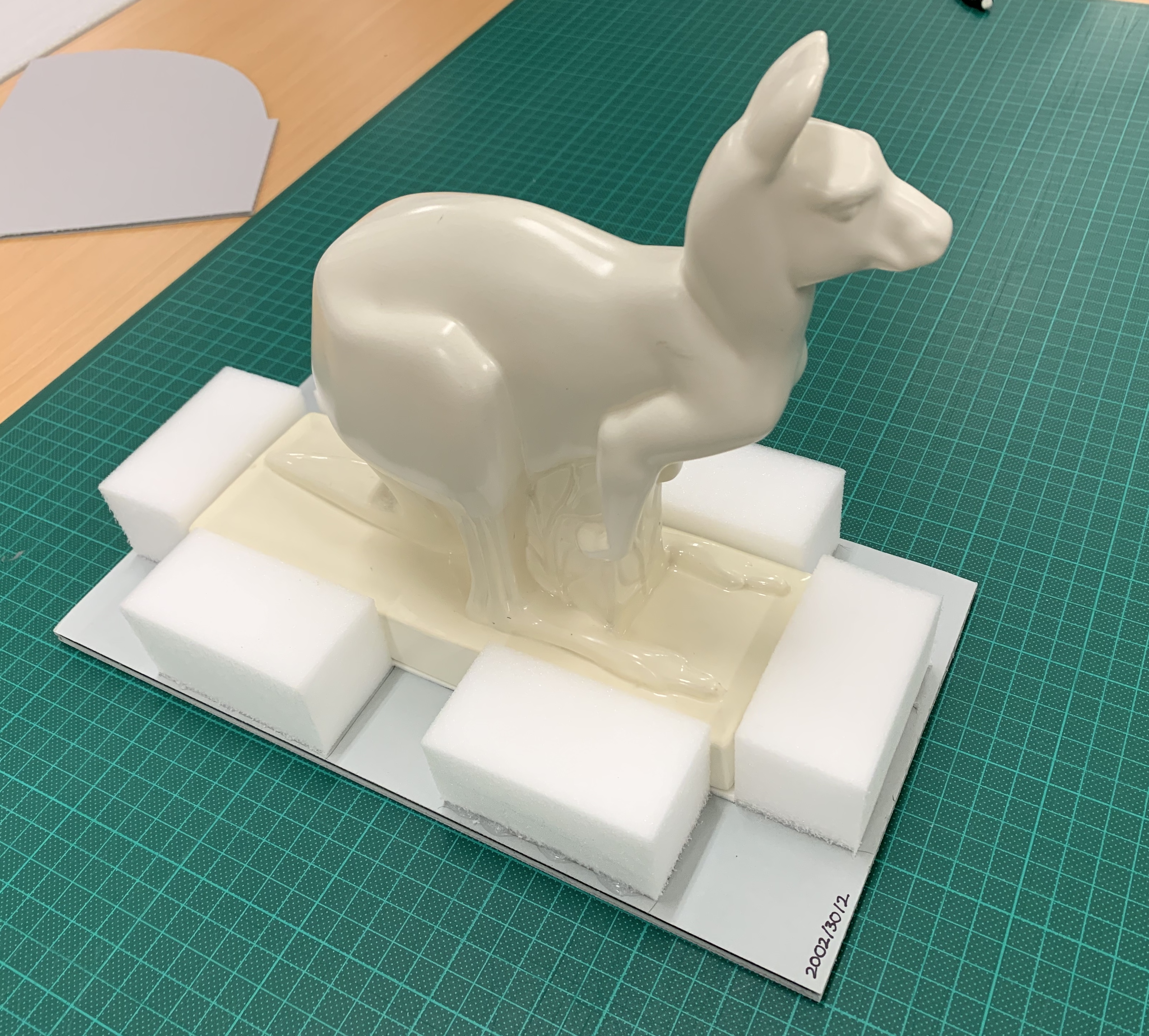 White earthenware kangaroo sitting on a stiff plastic base. The kangaroo figure itself has rectangular base around which have been glued six foam blocks – two each along the long sides of the base, and one each along the short sides.